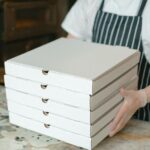 A pizza delivery worker holds stacked boxes in a restaurant kitchen, ready for delivery.